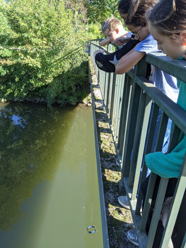 trois collégiens penchés sur la rembarde d'un pont regardent un fil et un poids qui descendent dans la rivière Nidda